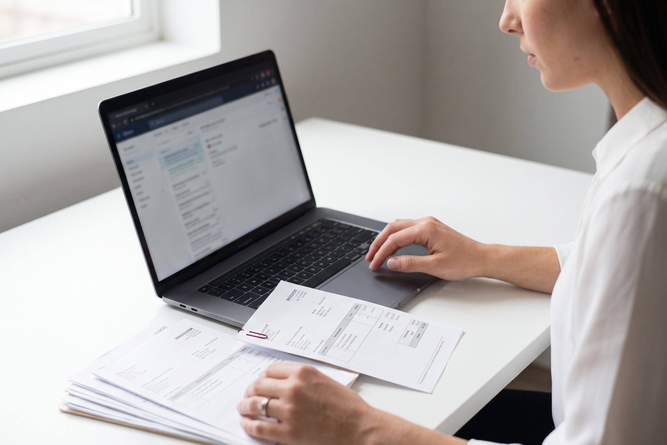 Hands checking a flagged invoice pulled from a stack beside a laptop